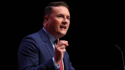 Wes Streeting against black backdrop, speaking and holding right hand up