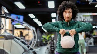 A woman in a green, zip-up workout jacket holds a white kettlebell at chest height. In the background, you can see a gym setting