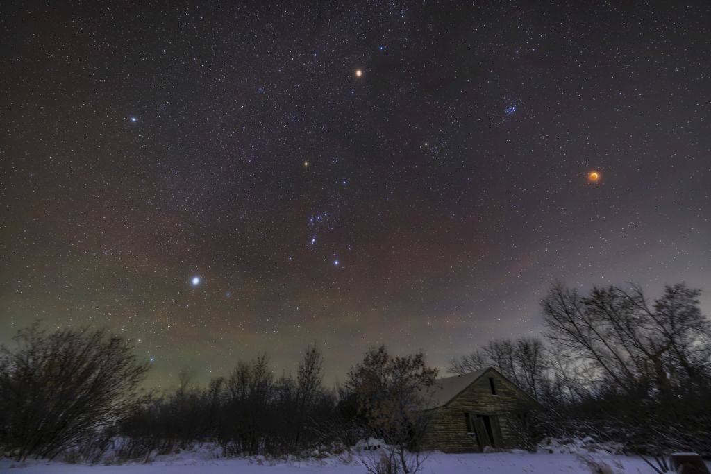 A series of bright stars dot a dark purple and green night sky over a wooden cabin surrounded by snow and bare trees.