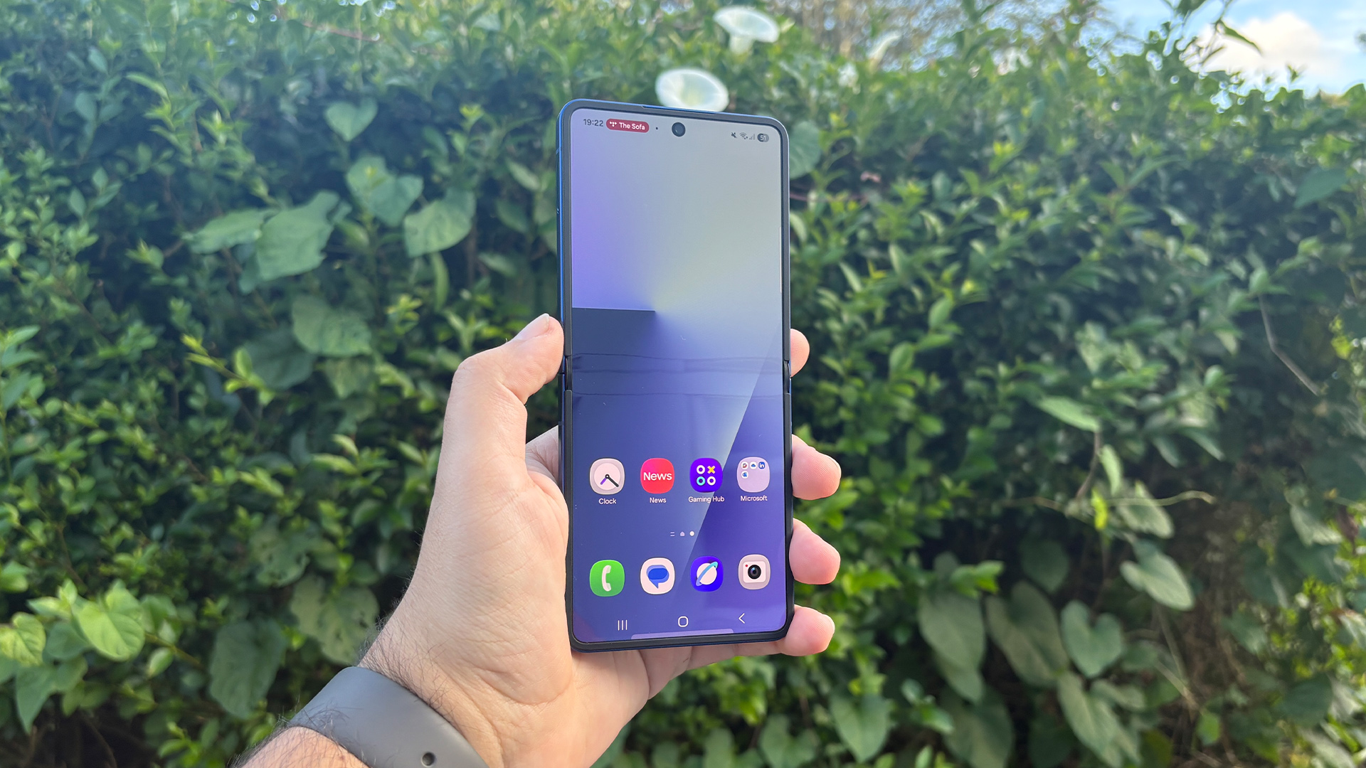 Samsung Galaxy Z Flip 7 smartphone held in hand in front of garden hedge showing apps on screen