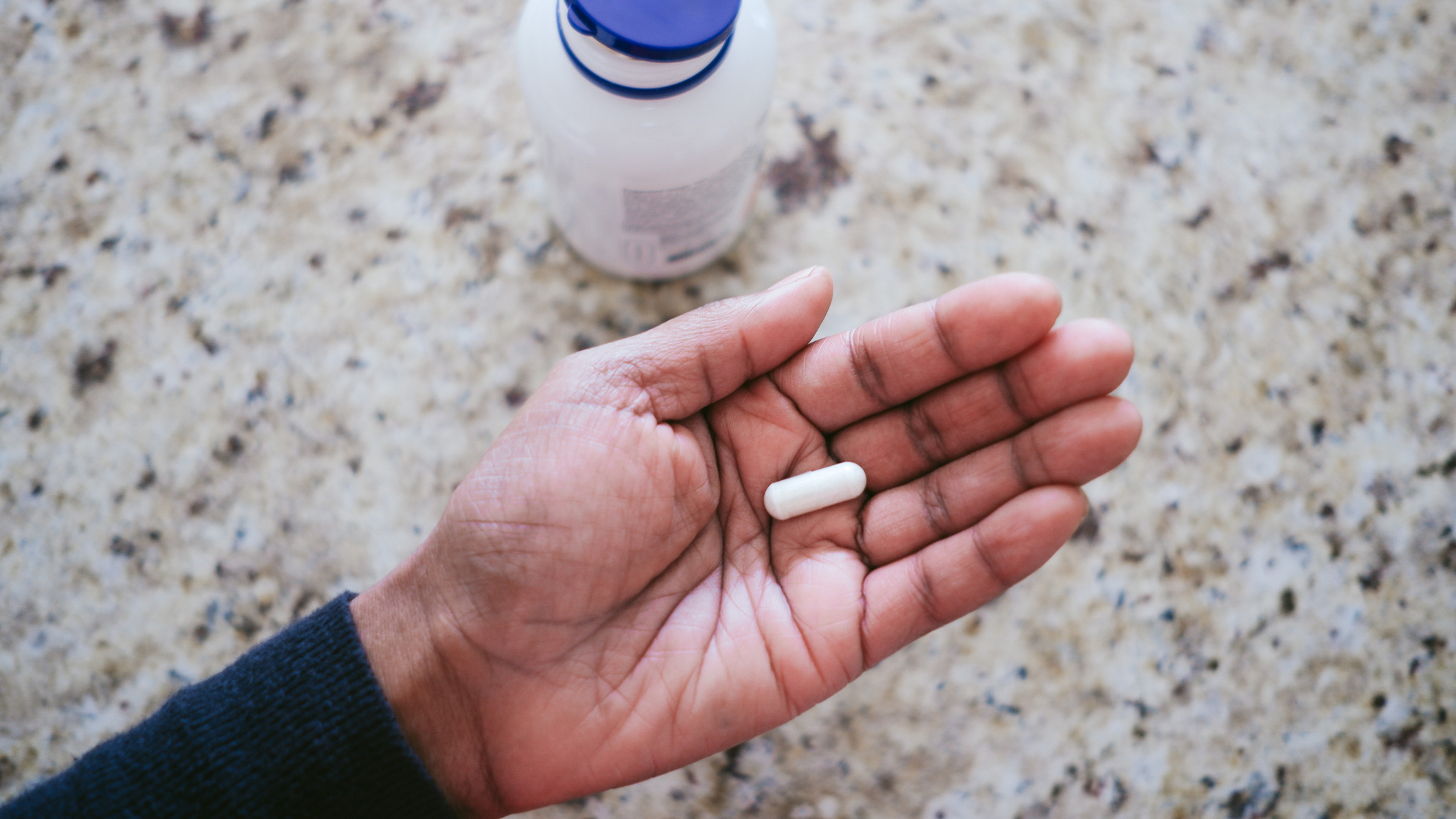 White pill held in palm of hand, next to a pill bottle with a blue lid