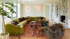 A white living room with a cool stone fireplace, a large olive green sofa on a patterned terracotta-colored rug with a curved wood coffee table and a pair of wood side stools near a plush brown sofa seater and a potted indoor tree