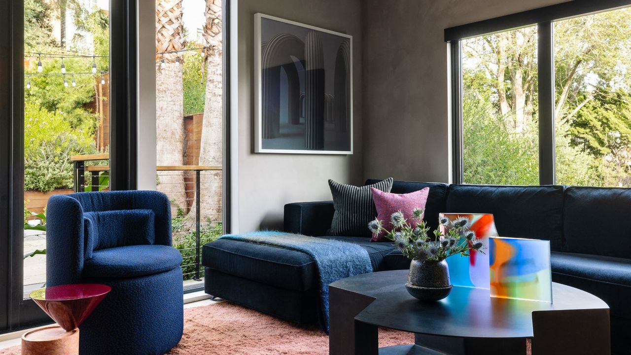 A living room with a blue couch and sofa chair, bright windows and a coffee table with colorful art and a vase