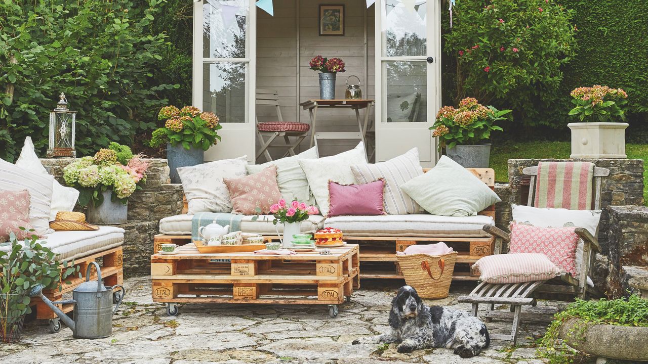 outdoor living with pallet furniture, recliner, pink and green colour scheme, watering can, potted planters, dog, shed in background 