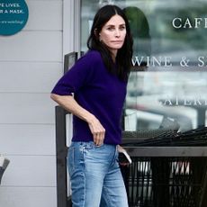 Courtney Cox wears grey trainers with slim-fitting blue jeans and a dark purple knit.