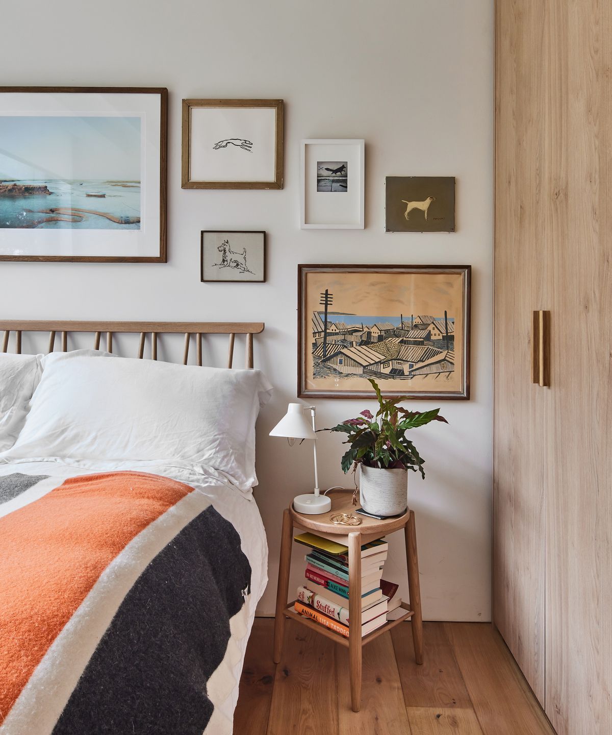 A wooden framed bed with bedside table filled with books with small fern plant and white reading lamp