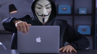 A hacker in a Guy Fawkes mask using an Apple MacBook.