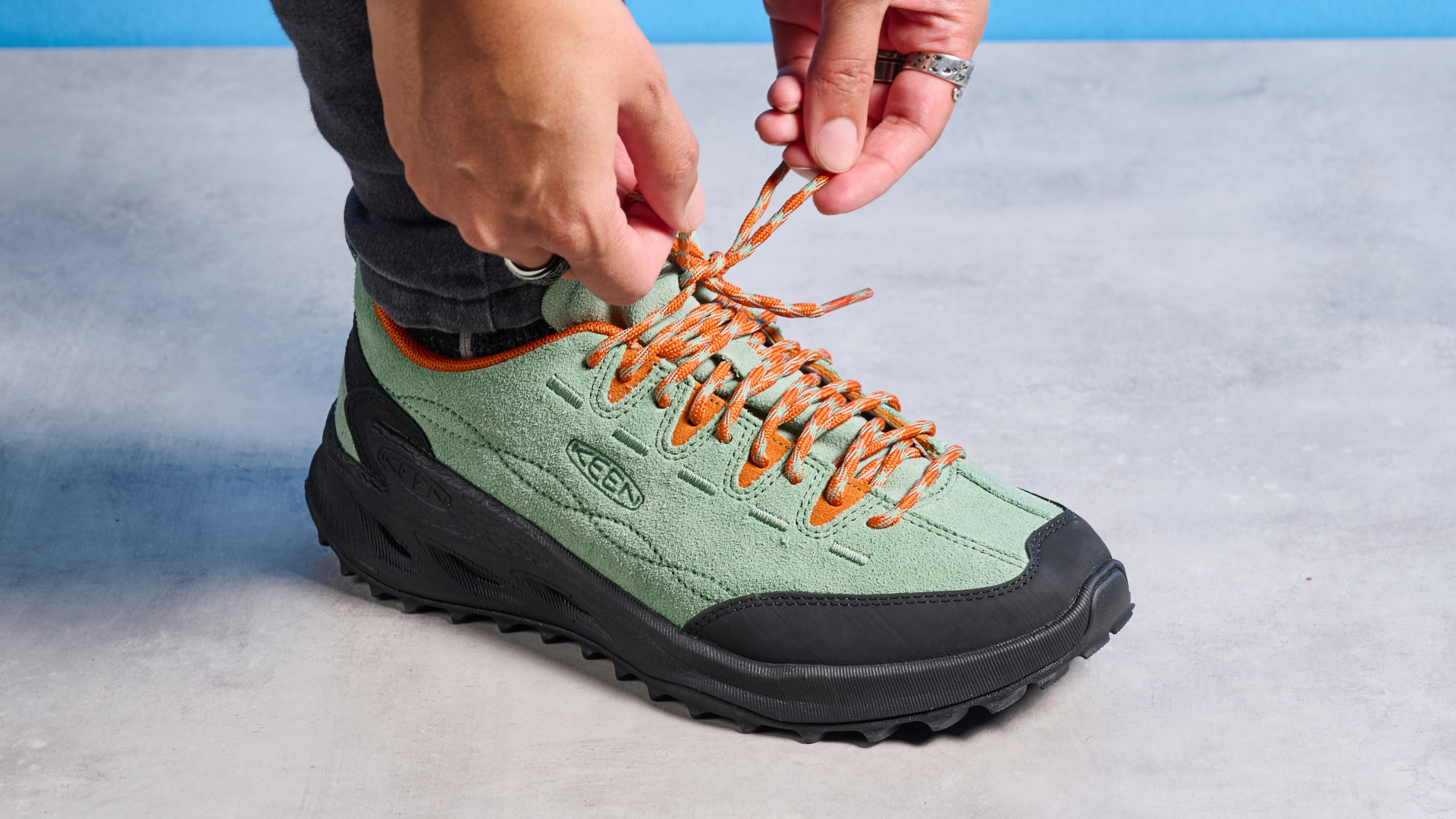 The Keen Jasper zionic hiking shoes in green/gold colorway are photographed against a blue Tom&#039;s Guide background and a stone floor, showing the lug sole and the leather upper with orange laces