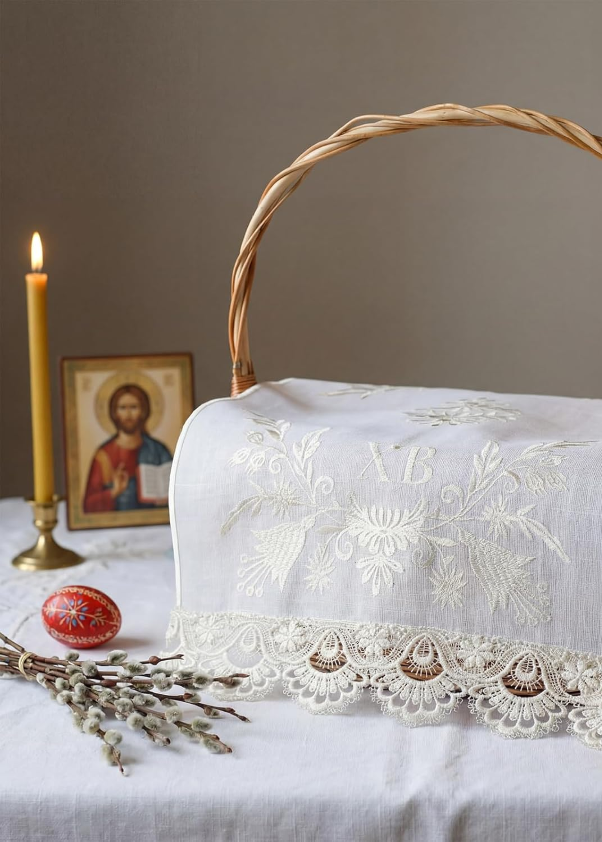 A basket with a lace cover, a bundle of twigs, a patterned egg, a candle, and an picture of Jesus Christ