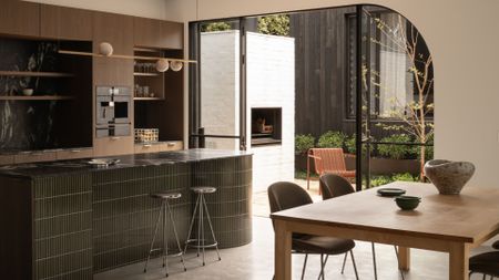 A kitchen and dining space with a green tiled island, chrome stools, a wood dining table, and a pendant light beside glass doors leading to an outdoor living room