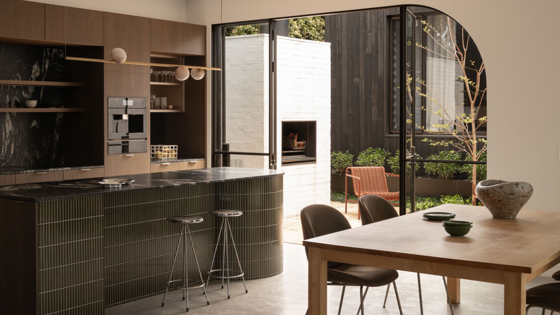 A kitchen and dining space with a green tiled island, chrome stools, a wood dining table, and a pendant light beside glass doors leading to an outdoor living room