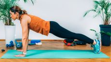 A woman performs a high plank exercise on an exercise mat. She is on the floor, supported by her tip toes and hands, while the rest of her body is held in a straight line. Behind her we see leafy plants, kettlebells and light dumbbells.