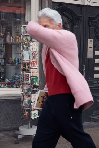 An elderly man walking in the street wearing a pink cardigan, a red shirt and dark blue pants.