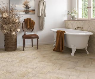 freestanding claw foot white bath in large bathroom with cream tiled floor, brown chair and large wicker pot with dried flowers