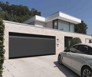 A modern new build home with flat roofs. The projecting single-storey ground floor is clad in stone and features a large section garage door from Hörmann. There is a sloping drive with a silver car parked on the drive.