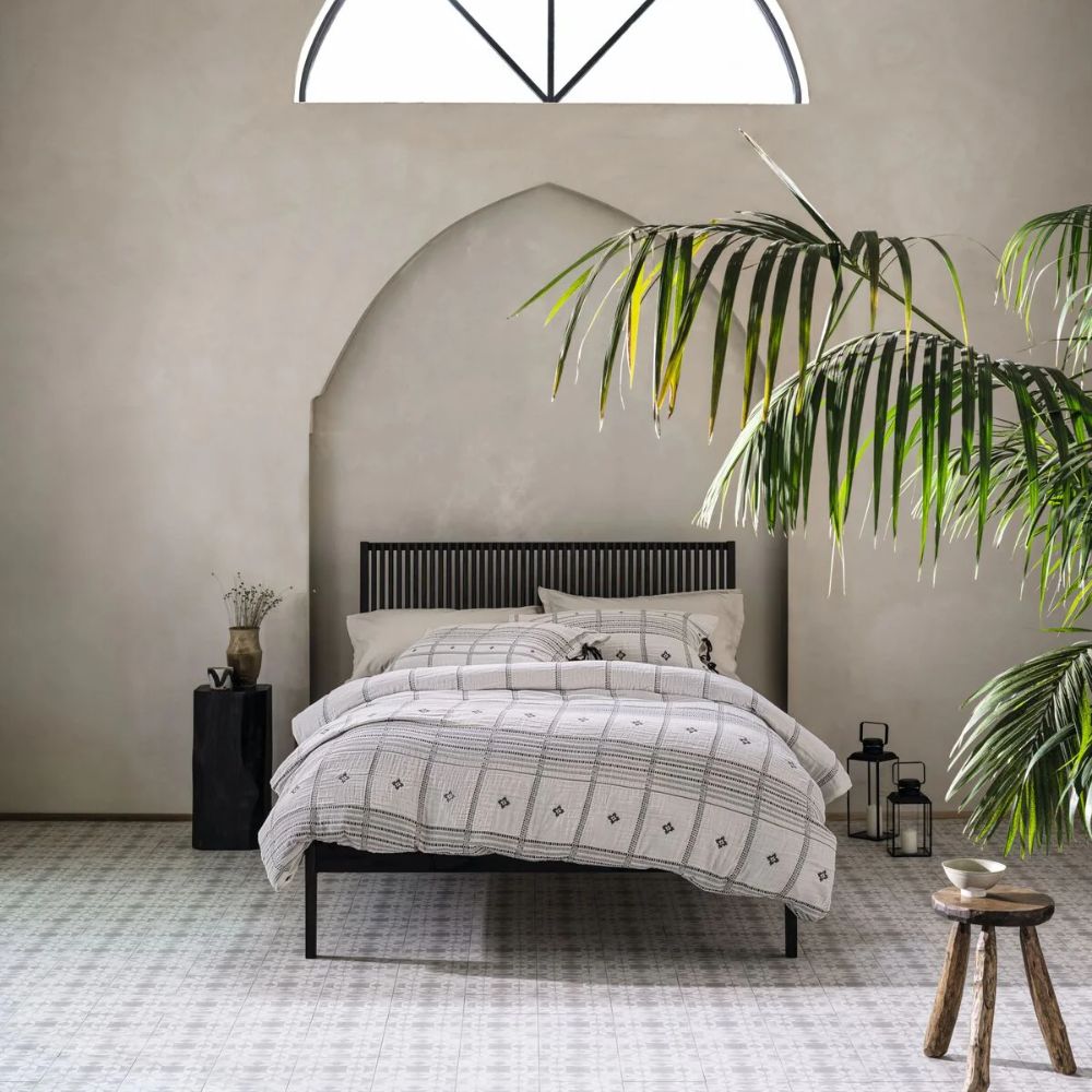 A serene, Moroccan-inspired bedroom with smooth sandy plaster walls, a pointed arch alcove and an arched metal-framed skylight above. A black slatted wood bed frame with a geometric patterned duvet sits on small white mosaic floor tiles, flanked by large tropical palm plants, a black lantern and a rustic wooden stool.