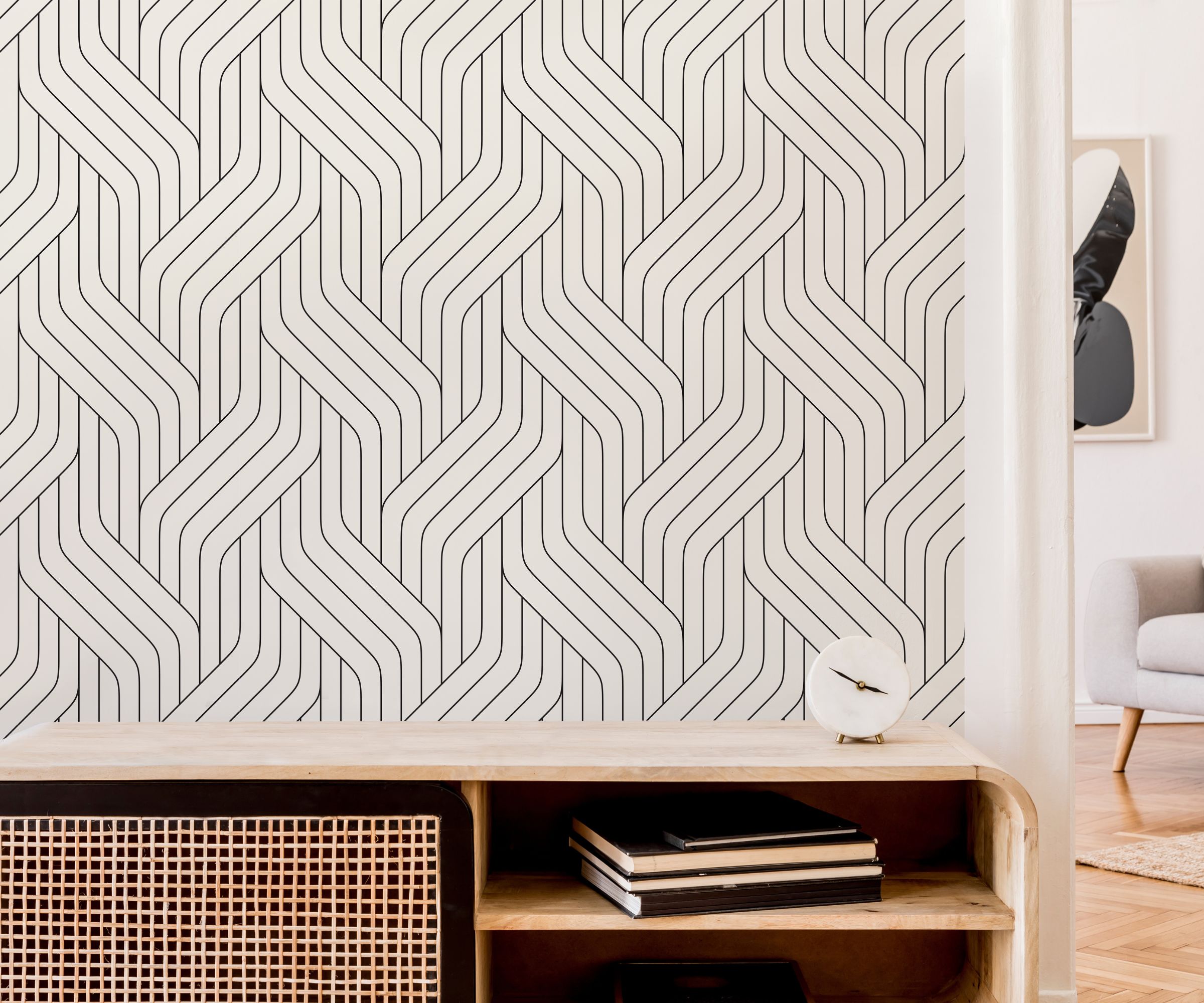 A wall covered in bold black-and-white geometric wallpaper with interlocking curved lines. In front sits a light wood sideboard with a rattan panel, stacked books, and a small round clock.