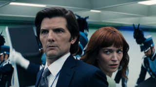 Mark and Helly standing back to back both looking on edge with a marching band behind them.