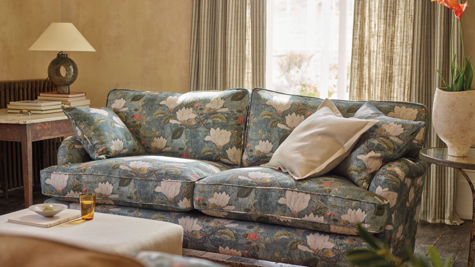 Cozy living room with blue sofa that has a large floral pattern, and floor length linen curtains letting in light where thick lined ones would not