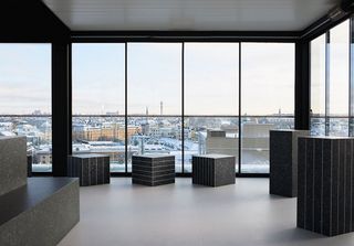 A design studio with grey vinyl flooring and floor to ceiling windows (giving of view of the city) featuring dark grey marble design box and rectangle shapes structures spread around the room