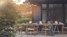 A wood panelled patio in front of a wood panelled home with a large tablescaped dining table, wooden chairs, and a large bush in the left foreground.