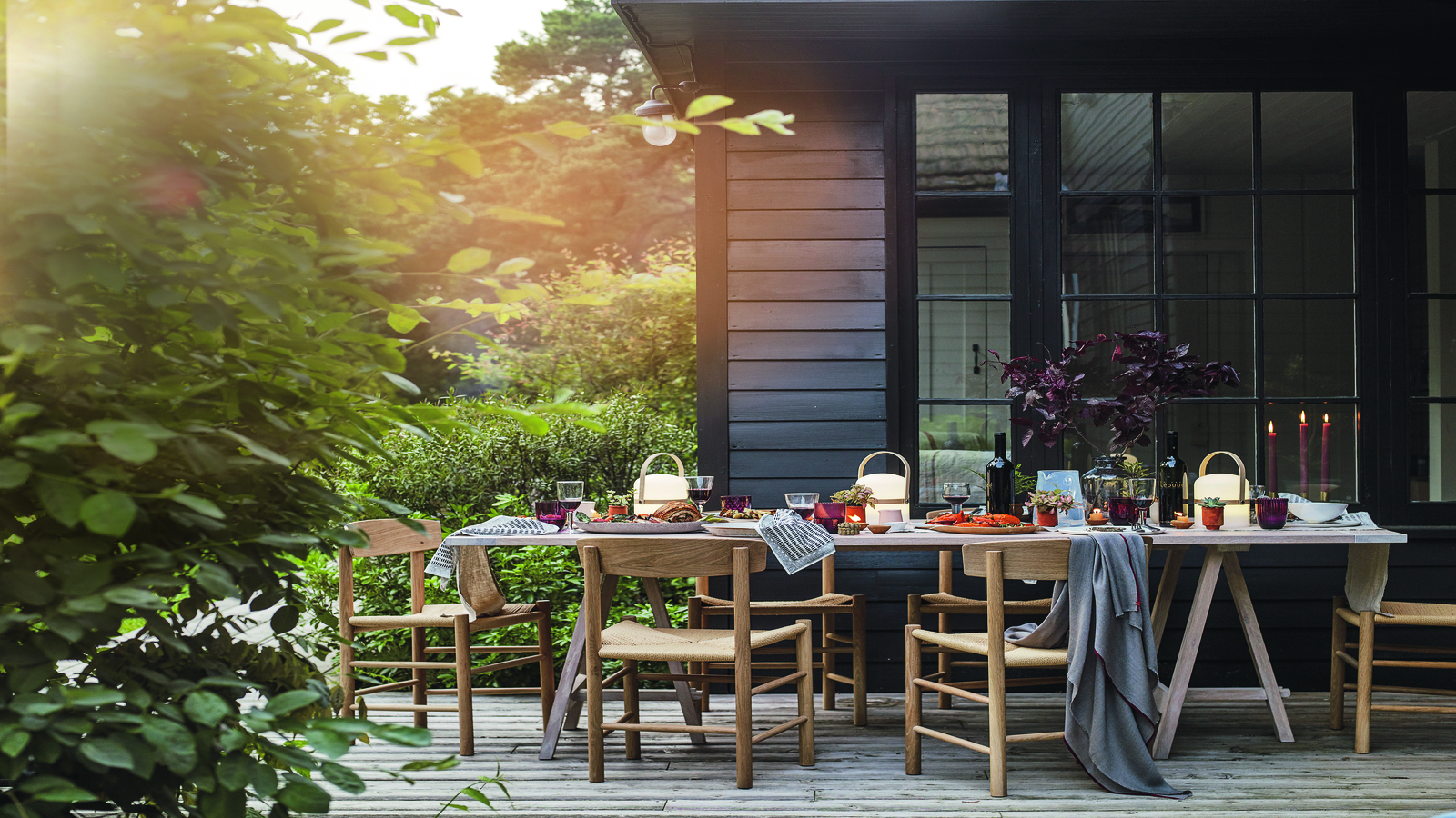 A wood panelled patio in front of a wood panelled home with a large tablescaped dining table, wooden chairs, and a large bush in the left foreground.