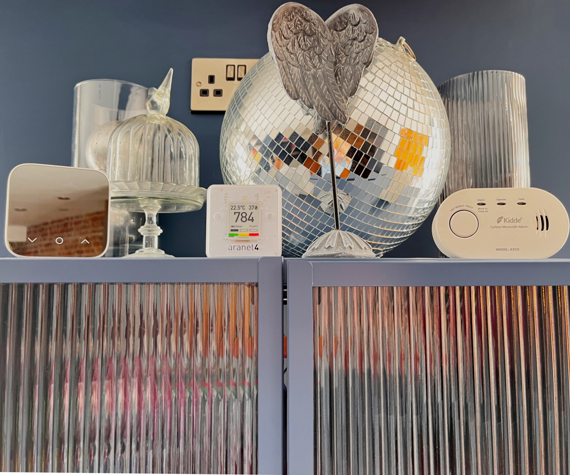 Aranet4 Home monitor on top of blue and reeded glass cabinet with mirrored disco ball, ornaments, carbon monoxide monitor and Hive thermostat