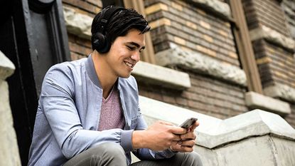 Best cheap headphones 2022, image shows Man wearing headphones on the street, holding phone and smiling