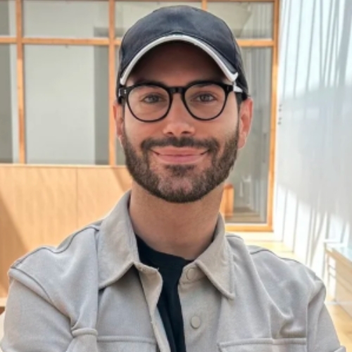 Image of a man in a black shirt and tan button-down with glasses and a black hat.