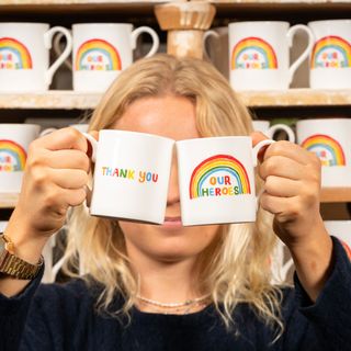 a blonde haired woman holding up two white mugs, one showing a rainbow with the words "our heroes" and the other one showing the other side with the words "thank you" written in rainbow colours