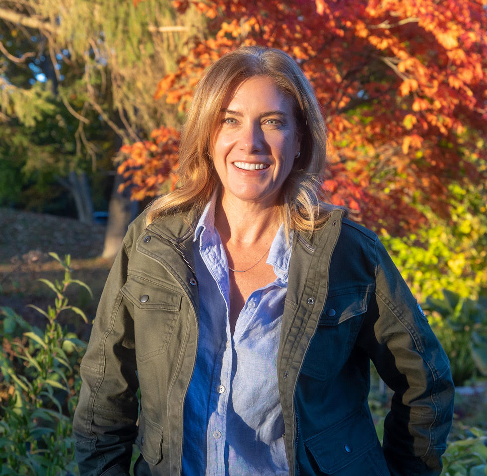 Woman with blonde hair in green jacket stood in front of trees