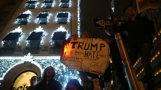 A group of people protesting outside an apartment building. One holds a sign that reads "Trump, make America hate again."