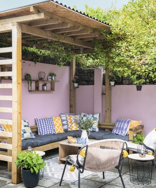 Wood pergola extension surrounding pink garden walls, with cozy lounge set-up, and blue and yellow scatter pillows.