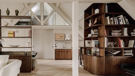 A refined attic apartment interior characterized by a warm, modernist aesthetic. The space is organized as an open-plan living area under a pitched roof, with exposed white-painted beams and triangular dormer windows that bring in soft natural light.