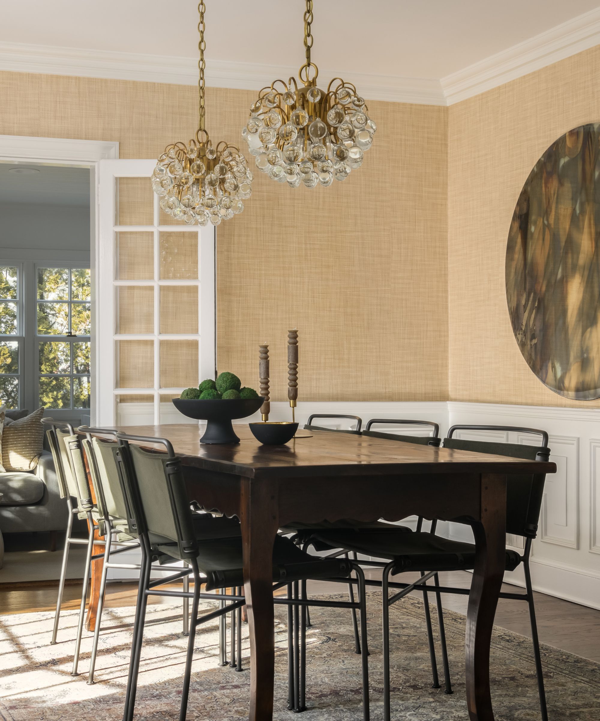A dining room with butter yellow wallpaper, white paneling on the lower walls, a large dark wood dining table with black chairs, and two matching brass and glass circular pendant lights.