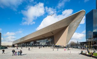 Daytime, front view of Rotterdam Central Station, paved walkway, visitors, trees, adjacent building, blue sky, white clouds