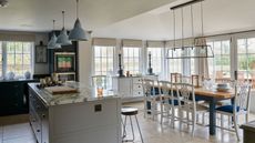 A white kitchen with pale beige tiles, a white island with marble countertop, and blue pendant lamps. To the right is a wooden dining table with white chairs. Large bright windows surround the walls.