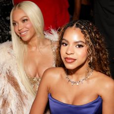 Blue Ivy wears a strapless gown and sits in front of her mom Beyonce, who has long blonde hair and is wearing a fur coat