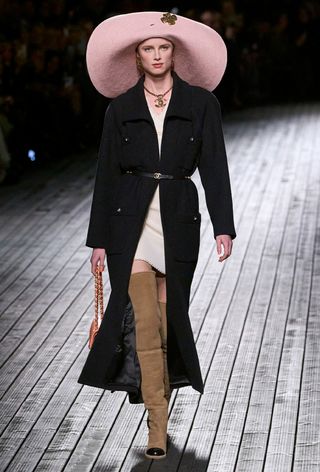 a photo showing some of the best thigh-high boots with a model walking the Chanel runway show wearing a pink floppy hat with a long black tweed cardigan styled over a white mini dress paired with brown thigh-high boots