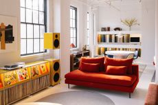 A red couch sits in a white-walled showroom alongside yellow speakers with a long cabinet of vinyl records.