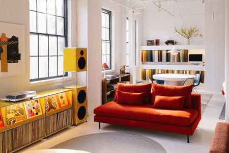A red couch sits in a white-walled showroom alongside yellow speakers with a long cabinet of vinyl records.