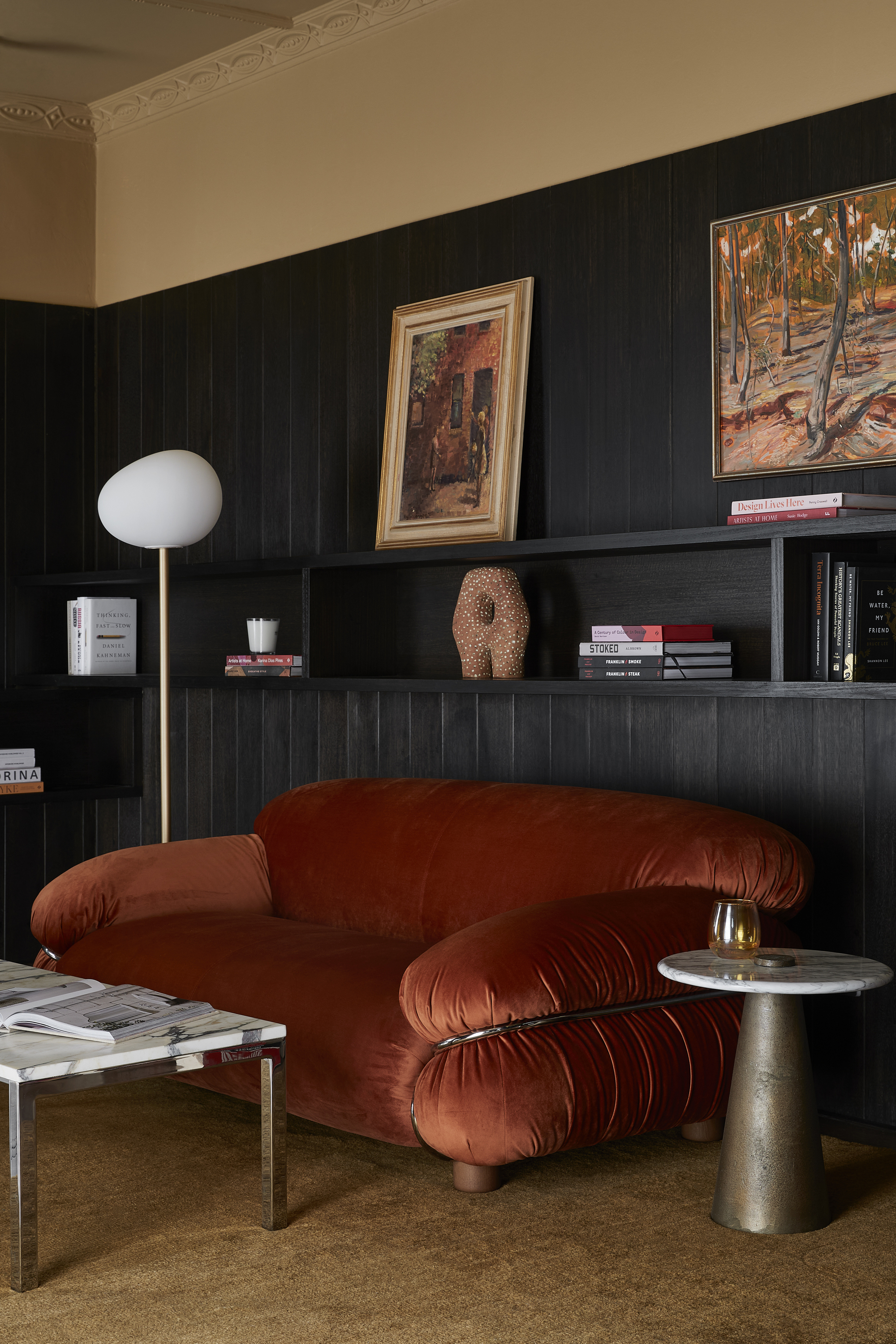 cozy living room with rust colored velvet sofa, caramel colored rug, dark black timber joinery with shelves decorated with accessories, and caramel color paint on the top wall and ceiling