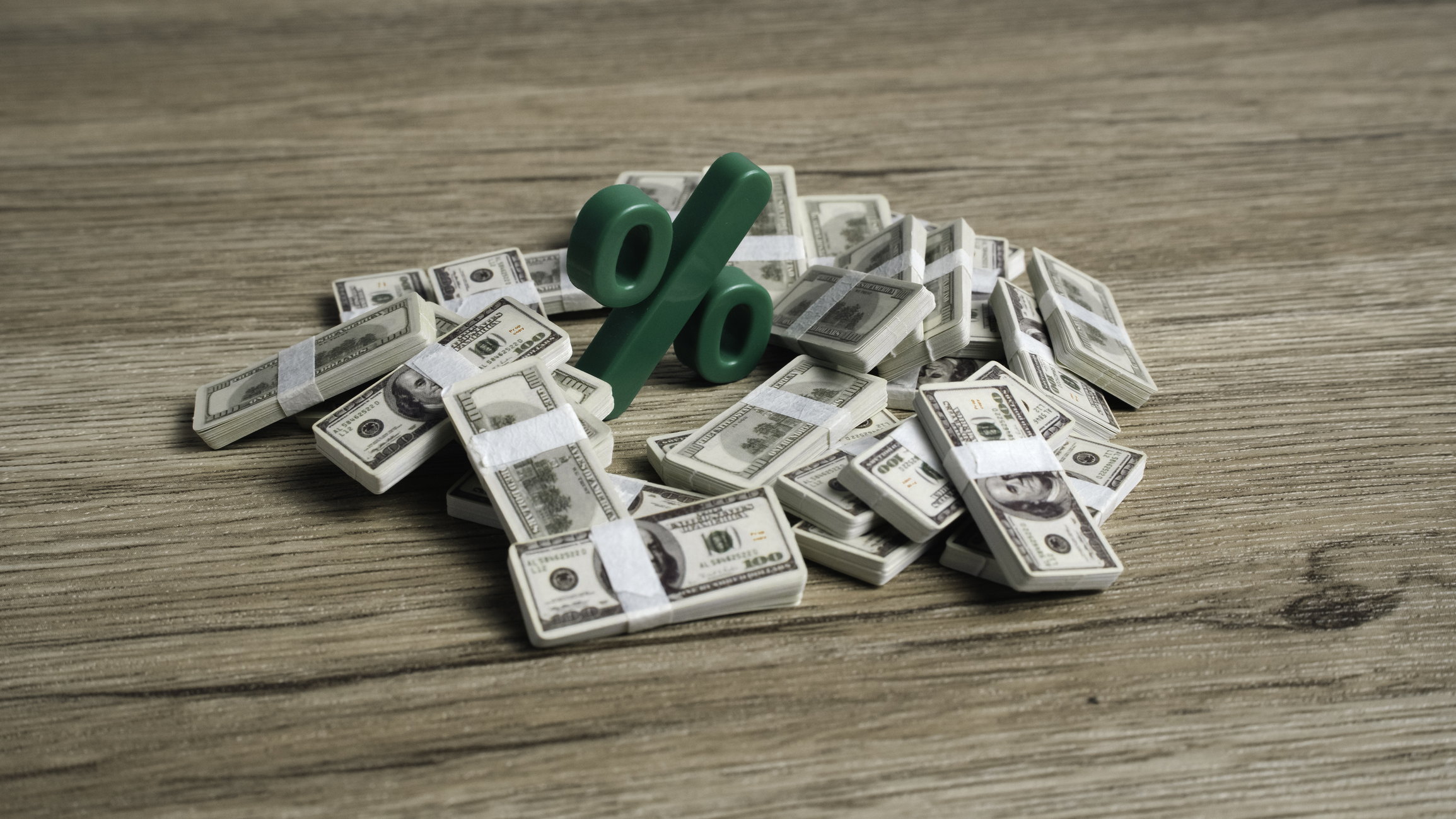 Green percentage symbol placed on top of a large pile of hundred dollar bill bundles on a wooden table.