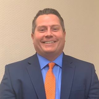 A head shot of Glenn Robbins he is wearing a suit with a blue shirt and orange tie.