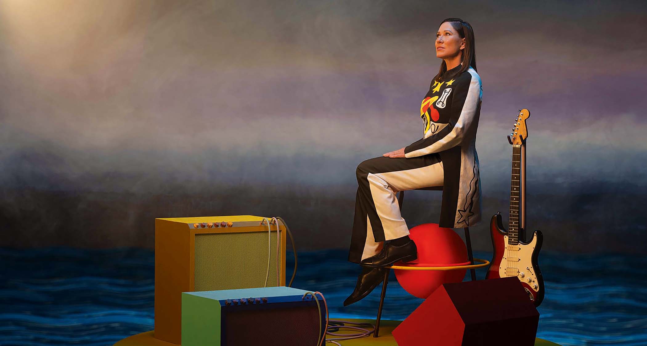 A stylized press shot of Kim Deal to accompany her new album Nobody Loves you more. She sits hight on a chair in front of a combo amp. her Fender Strat sitting in its stand.