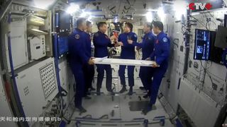 six astronauts in blue flight suits float aboard a space station with white walls.