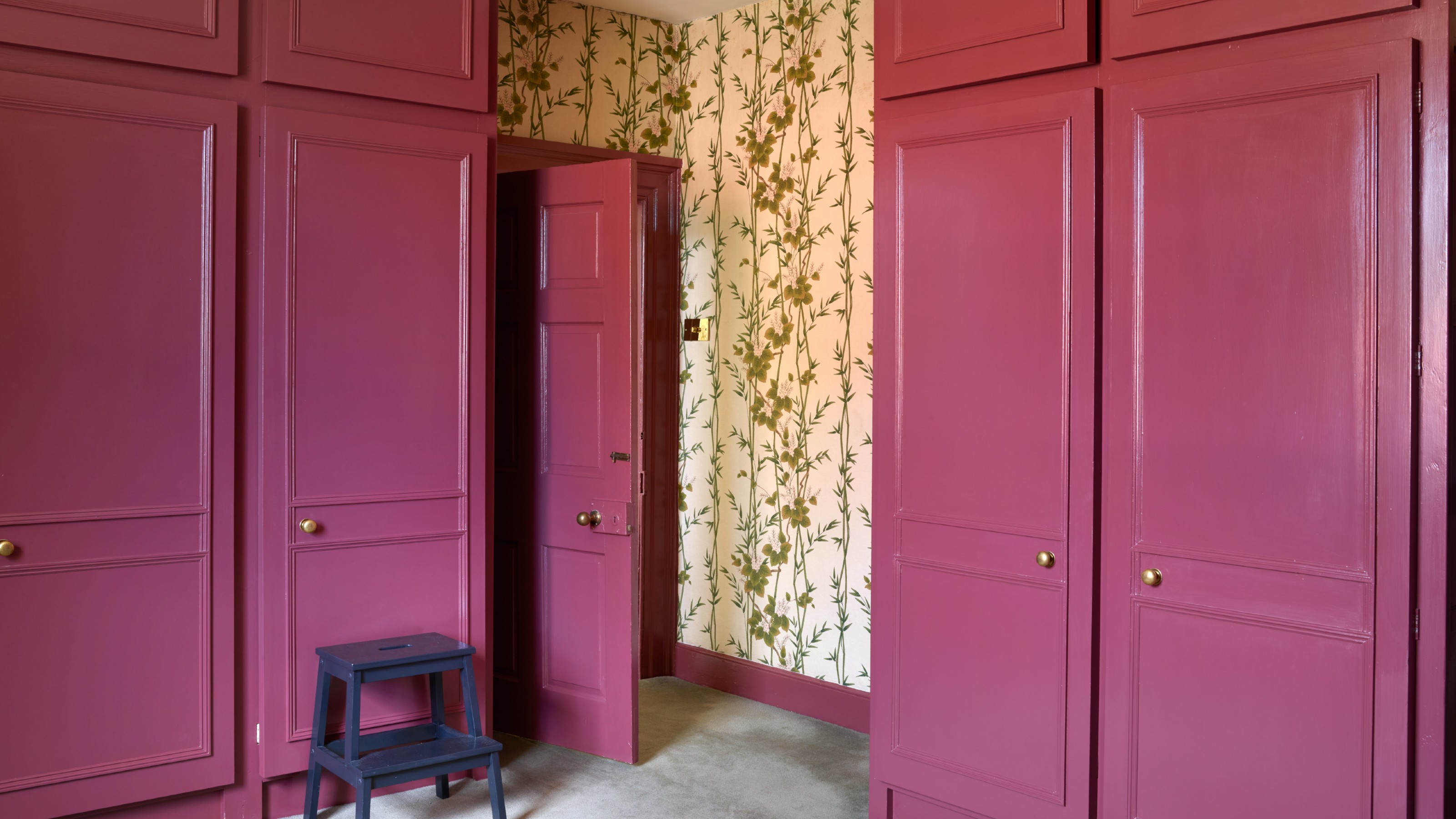 A room with wardrobes painted in a dark pink glossy paint set against a floral wallpaper on the walls