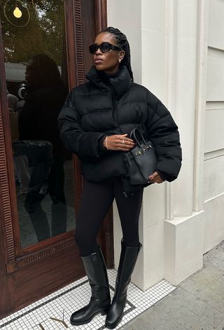 a roundup of the best puffer jackets shown in a photo of a woman wearing a black puffer coat over a black turtleneck with black leggings and knee-high boots