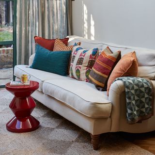 classic neutral sofa covered in bold patterned and coloured cushions with a red gloss footstool