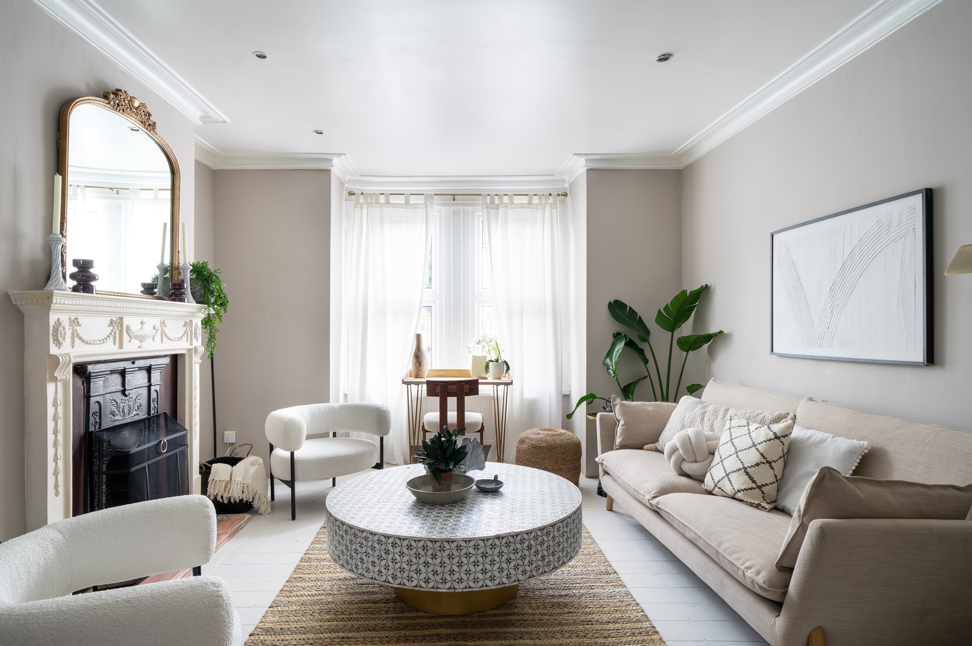 modern living room with beige walls, sheer curtains, round mosaic coffee table, beige sofa, ornate fireplace with mirror above and boucle armchairs and jute rug